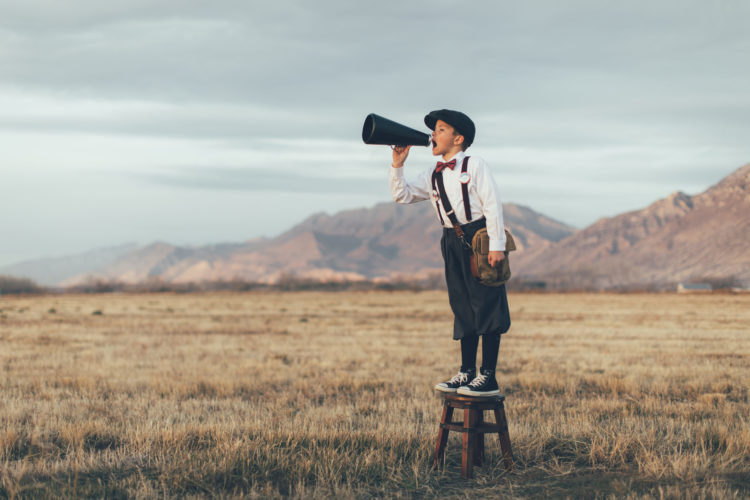 A news boy dressed in vintage knickers and newsboy hat stands yelling through a megaphone in the middle of a field in Utah, USA. He is trying to sell you what your business needs.